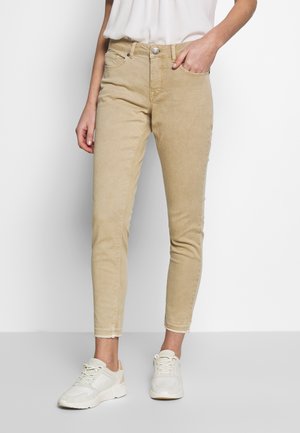 Beige Slim-Fit-Jeans mit ausgefransten Säumen, getragen von einer Person in weißen Sneakers und einem in die Hose gesteckten weißen Hemd, eine Hand in der Tasche.