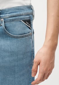 Κοντινό πλάνο του χεριού ενός ατόμου δίπλα σε μπλε τζιν με μικρή μαύρη υφασμάτινη ετικέτα που γράφει "REPLAY" στην άκρη της μπροστινής τσέπης.