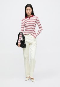 Czerwona i biała pasiasta koszulka polo z długim rękawem, zestawiona z wysokim stanem kremowych spodni. Modelka trzyma czarną torbę na ramię.
