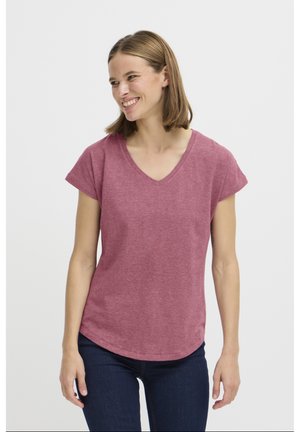Femme aux cheveux mi-longs souriante, portant un t-shirt rose à manches courtes et col en V, et un jean bleu foncé sur un fond blanc uni.