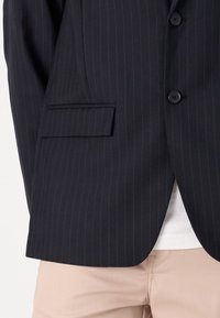 Marineblaues Nadelstreifen-Sakko mit einer Brusttasche, zwei Knöpfen und einer glatten Textur. Darunter ist ein weißes Hemd teilweise sichtbar.
