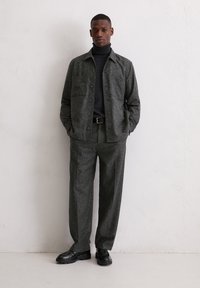 Graue, strukturierte Jacke mit passender Hose, die vorne mit Knöpfen verschlossen wird und zwei Brusttaschen hat. Schwarzer Rollkragenpullover und schwarze Schuhe.