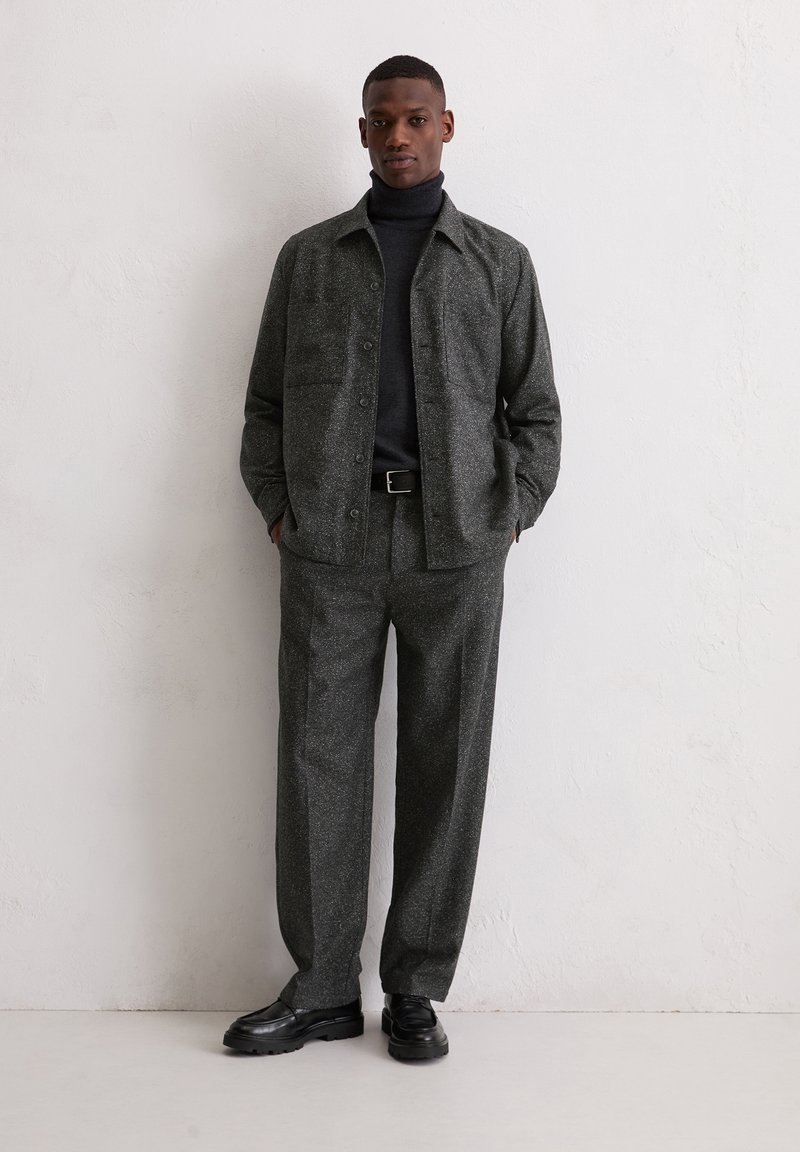 Graue, strukturierte Jacke mit passender Hose, die vorne mit Knöpfen verschlossen wird und zwei Brusttaschen hat. Schwarzer Rollkragenpullover und schwarze Schuhe.