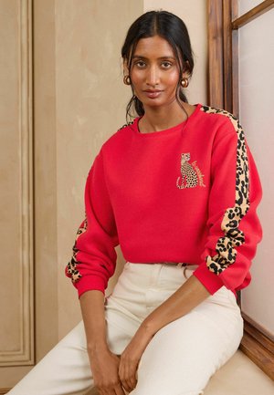 Frau trägt ein rotes Sweatshirt mit Leopardenstreifen an den Ärmeln und einer kleinen Leoparden-Grafik, kombiniert mit weißen hoch taillierten Hosen.