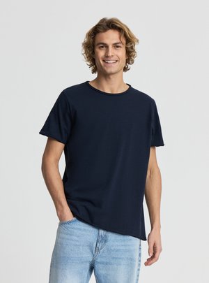 Giovane sorridente con capelli ricci che indossa una maglietta blu scuro semplice e jeans azzurri, in piedi davanti a uno sfondo neutro.