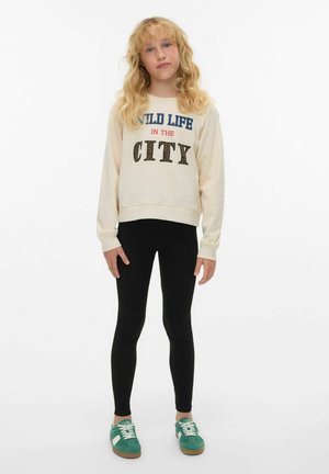 Krämfärgad sweatshirt med grafiken "WILD LIFE IN THE CITY", svarta leggings och teal-färgade sneakers. Slätt tyg, avslappnad passform, ribbade muddar.