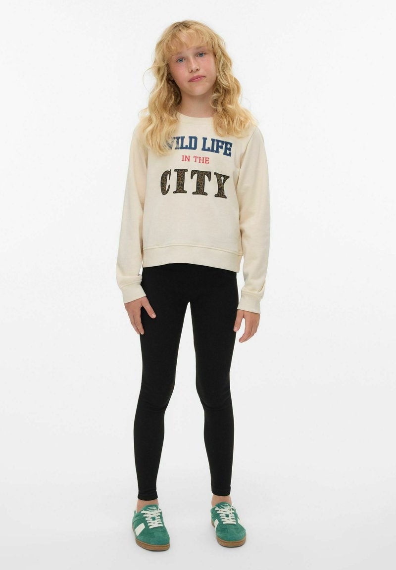 Krämfärgad sweatshirt med grafiken "WILD LIFE IN THE CITY", svarta leggings och teal-färgade sneakers. Slätt tyg, avslappnad passform, ribbade muddar.