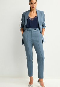 Hellblau maßgeschneiderter Blazer und passende Slim-Fit-Hosen, ergänzt durch ein navyfarbenes Spitzen-Cami und silberne hochhackige Schuhe.