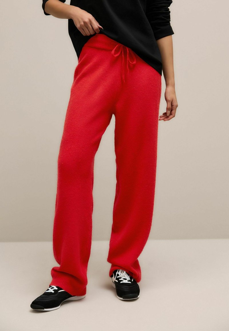 Pantaloni din fleece roșii, moi, cu o croială lejeră, talie cu șnur și mansete ribbate, purtați cu pantofi sport negri cu accente albe.