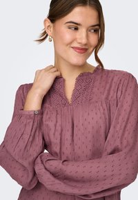 Νεαρή γυναίκα με καστανά μαλλιά, που φοράει χρυσούς κρίκους στα αυτιά και μια μακρυμάνικη, υφασμάτινη μωβ μπλούζα με λεπτομέρειες από δαντέλα, χαμογελώντας και κοιτάζοντας πλάγια.