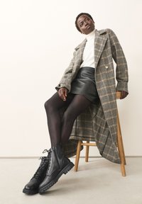 Geruite lange jas, witte coltrui, zwarte leren rok, ondoorzichtige panty's en zwarte veterlaarzen met dikke zolen, zittend op een houten kruk.