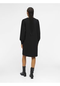 Schwarzes Kleid mit Puffärmeln, knielang geschnitten und gerafften Rückendetails. Matte Textur kombiniert mit knielangen, schwarzen, höchsten Stiefeln.