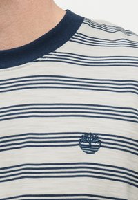 Κοντινό πλάνο λευκού και ναυτικού μπλε ριγέ πουκάμισου με ναυτικό μπλε λαιμόκοψη και κεντημένο λογότυπο δέντρου στο στήθος.