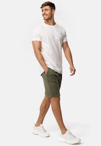 Camiseta blanca de cuello redondo, pantalones cortos verdes oliva y zapatillas blancas. Los pantalones cortos tienen bolsillos laterales y un corte recto. Marca mínima en la camiseta.