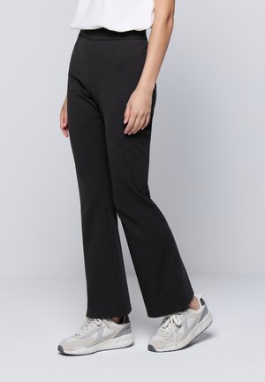 Pantalons noirs évasés à taille haute, fabriqués à partir d'un tissu lisse et extensible. Portés avec des baskets de sport grises et blanches.