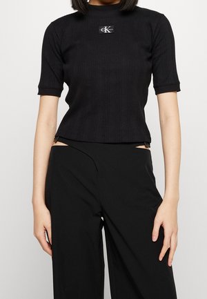Woman wearing a black ribbed short-sleeve top with Calvin Klein logo and high-waisted black pants with cutout waist details.