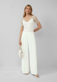 Haut à manches en dentelle blanche associé à un pantalon large taille haute de couleur pâle. Accentué par un petit sac à main blanc et des sandales à lanières.