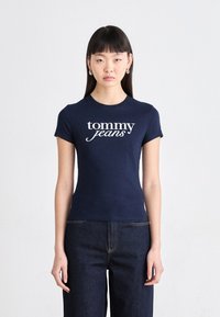 Tamsiai mėlynas medvilninis marškinėlis trumpomis rankovėmis su baltu "tommy jeans" logotipu švelniu šriftu, derinamas su tamsiai mėlynomis džinsinėmis kelnėmis.