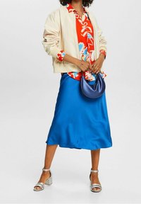 Veste bomber crème, blouse à boutons rouge à motif floral, jupe en satin bleu, sandales à talons argentées et sac à main bleu marine. Textures lisses et couleurs vibrantes.