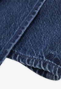 Dongeristoff i mørkeblått med en teksturert overflate og kontrasterende sømmer. Har en brettet kant og subtile broderidetaljer.