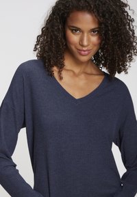 Woman with curly hair wearing a dark blue ribbed V-neck long-sleeve top, smiling slightly against a plain background.