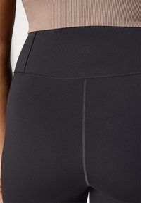 Leggings noirs taille haute avec une texture lisse, présentant un logo en relief et des coutures contrastées. Fabriqués en tissu extensible.
