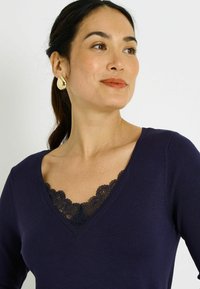 Suéter de punto azul marino con escote en V adornado con ribete de encaje negro, textura suave y mangas largas, acentuado por un delicado patrón floral.