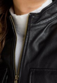 Black leather jacket with a gold zipper, featuring a white fleece lining and two front pockets. Smooth texture and structured design.