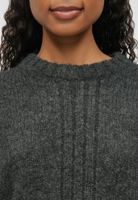 Σκούρο γκρίζο πλεκτό πουλόβερ με ψηλό λαιμό, το οποίο διαθέτει υφή με κάθετες ραβδώσεις και απαλό, φουντωτό ανάγλυφο.