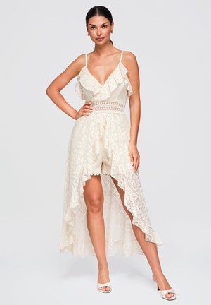 Femme portant une robe blanche en dentelle sans manches avec des volants et un ourlet asymétrique, debout avec une main sur la hanche et des talons blancs.