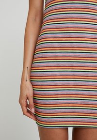 Tatouage de bras avec flèche le long du poignet, à côté d'une robe en tissu côtelé à rayures horizontales multicolores contre un fond gris clair.