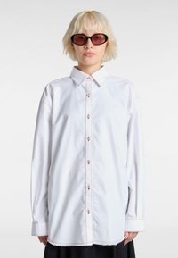 White button-up shirt with a classic collar, visible red stitching, and long sleeves. The hem is slightly frayed, creating a relaxed fit.