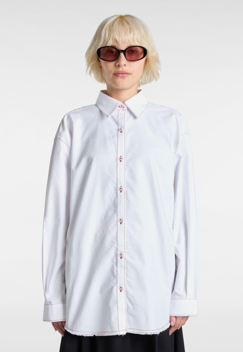 White button-up shirt with a classic collar, visible red stitching, and long sleeves. The hem is slightly frayed, creating a relaxed fit.