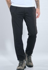 Mann trägt schwarze enge Hose und graue Schnürstiefel, steht mit den Händen in den Taschen vor hellem Hintergrund.