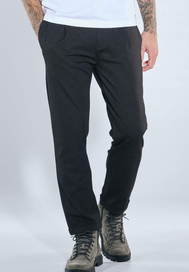 Mann trägt schwarze enge Hose und graue Schnürstiefel, steht mit den Händen in den Taschen vor hellem Hintergrund.