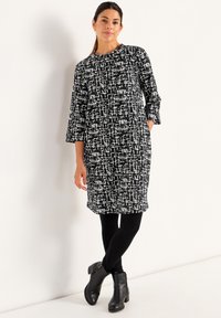 Vrouw die tegen een witte muur staat, gekleed in een zwart-wit gestreepte jurk, zwarte leggings en zwarte enkellaarsjes.