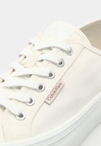 Sneaker bianco in tela con superficie testurizzata, occhielli rotondi in argento, lacci bianchi e un piccolo tag Calvin Klein vicino alla caviglia.