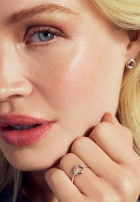 Silverfärgade stuvar och en matchande ring med cirkulär design. Slät textur, polerad yta, bärs på en hand som rör vid ansiktet.