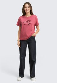 Roze katoenen T-shirt met een bloem- en vogelmotief, gecombineerd met donkerblauwe denim wijde broekspijpen en witte sneakers.