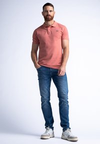 Polo shirt corail, manches courtes, couleur unie ; associé à un jean en denim bleu et des baskets blanches avec des accents gris.