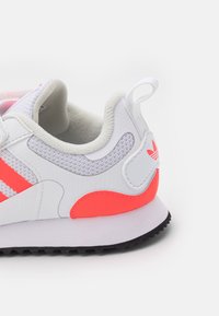 Białe sportowe buty z szarym siateczkowym wstawieniem, różowymi akcentami i textured gumową podeszwą. Posiadają uchwyt oraz logo w trzy paski.