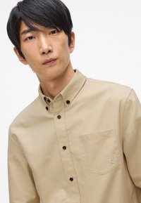 Beige knoopshirt met een puntkraag, een borstzak, zwarte knopen en subtiele geborduurde details op de borstzak.