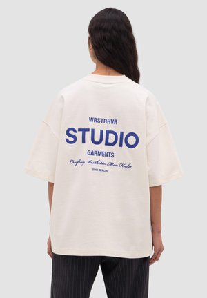 Person with long dark hair wearing a white oversized T-shirt with blue text "WRSTBHVR STUDIO GARMENTS" on back and black pinstripe pants.