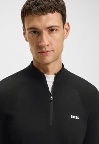 Schwarzer Reißverschluss-Pullover mit strukturierten Schulterpaneelen, hohem Kragen und einem weißen Logo auf der Brust. Der Stoff wirkt glatt und enganliegend.