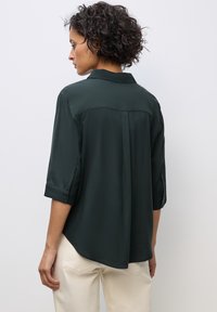 Mörkgrön, silkeslen blus med krage, uppvikta ärmar och en lätt svans. Har en jämn textur och minimalistisk design.