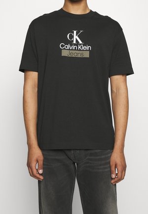 T-shirt noir à manches courtes avec le logo "cK Calvin Klein Jeans" en blanc et beige porté par une personne debout portant un jean foncé.