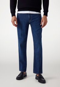 Jeans in denim blu scuro con vestibilità dritta, cuciture accentuate e un classico design a cinque tasche. Abbinati a mocassini in pelle nera.