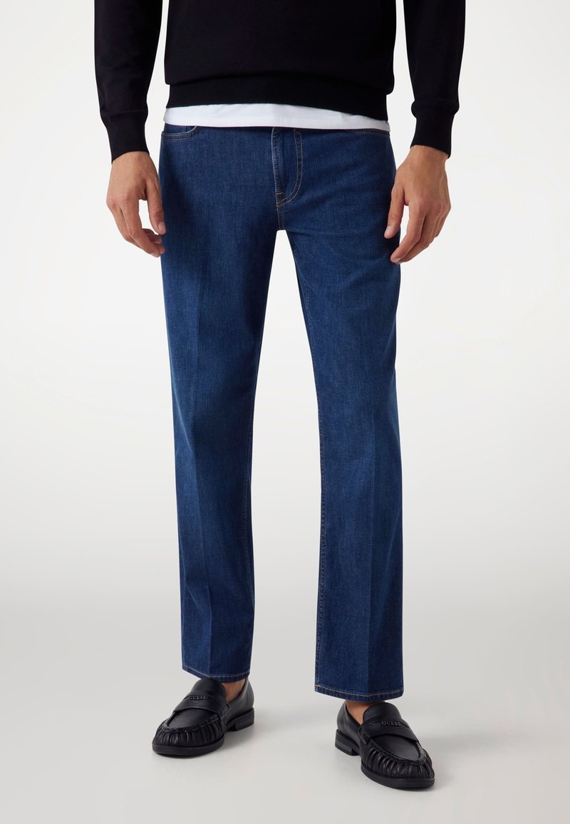 Jeans in denim blu scuro con vestibilità dritta, cuciture accentuate e un classico design a cinque tasche. Abbinati a mocassini in pelle nera.