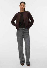 Giubbotto bomber in satin viola scuro con colletto rotondo, indossato sopra un top in maglia nera strutturata, abbinato a jeans grigi chiari a gamba dritta.