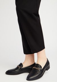 Mocassins en cuir noir avec une finition texturée, semelle plate et accent décoratif en chaîne dorée. Associés à un pantalon noir court.